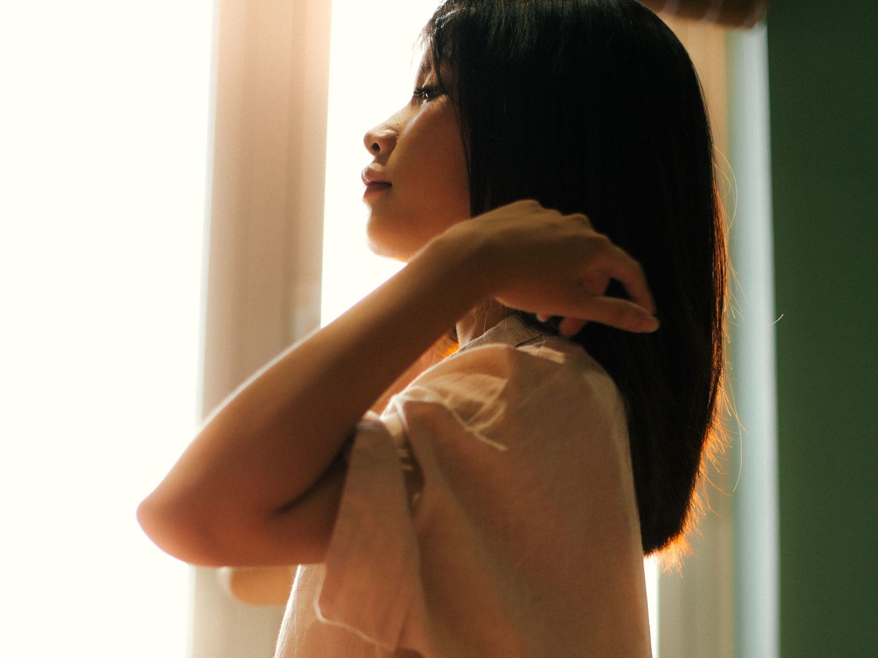 A woman with shoulder-length dark hair stands by a sunlit window in the morning, tucking her hair behind her shoulder